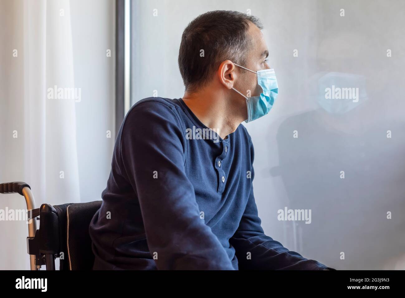 Young man with medical protective mask looking out the window. Man in isolation at home. Focus on his face. Stock Photo