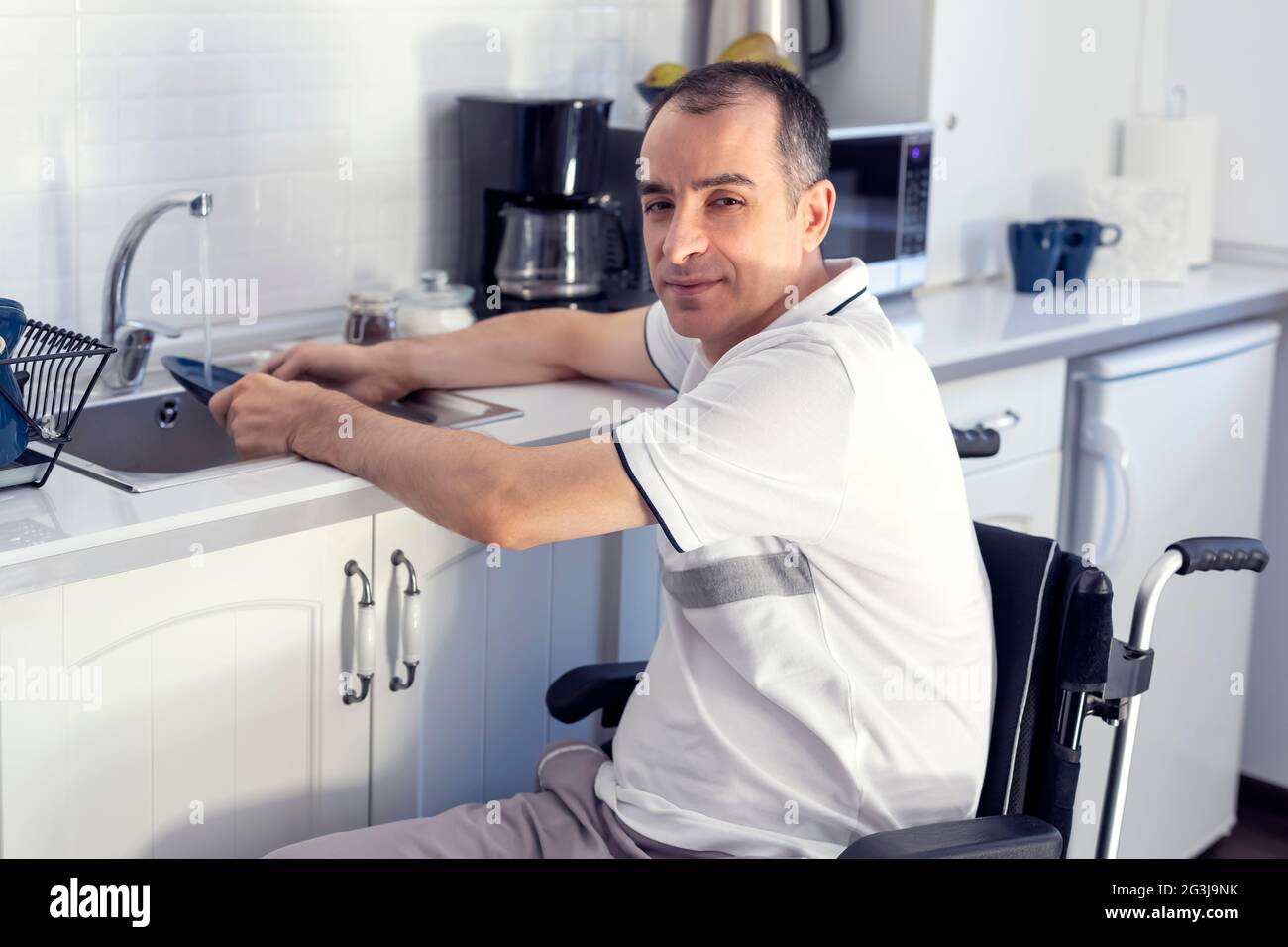 Young man Disabled Man On Wheelchair Washing Dishes. Smiling Young Handicapped Man Sitting On Wheelchair In Kitchen. Focus on his face. Stock Photo