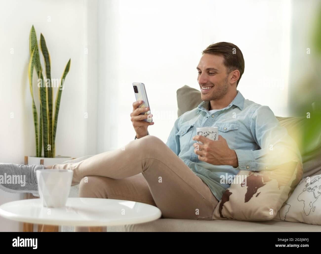 young man smiling, holding and looking at his smartphone, relaxed on the sofa at home drinking a coffee Stock Photo