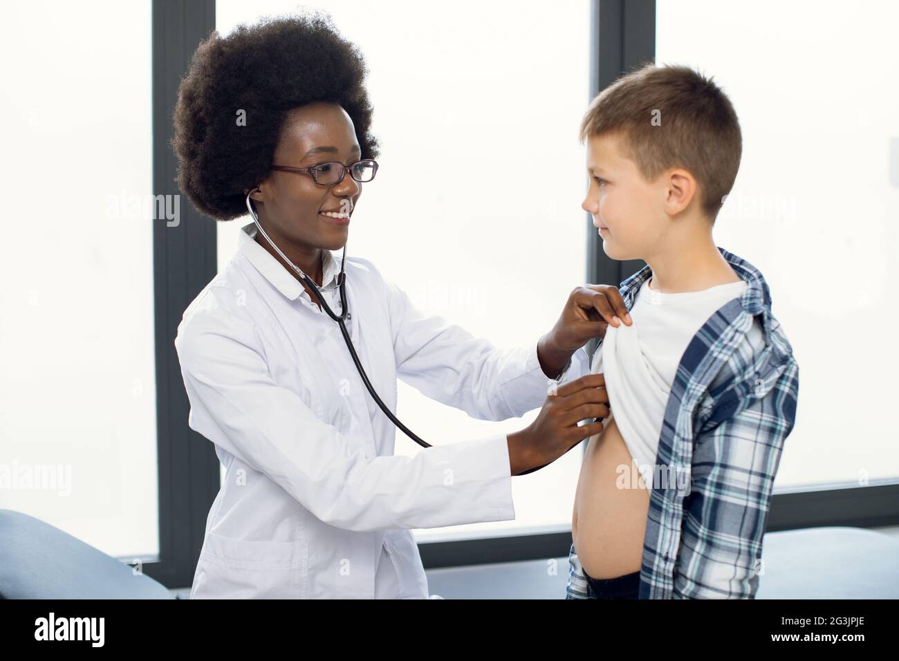 Cute likable Caucasian teen boy and joyful African woman doctor, during doctors checkup. Pediatrist examinates young patient's heartbeat and lungs with stethoscope Stock Photo