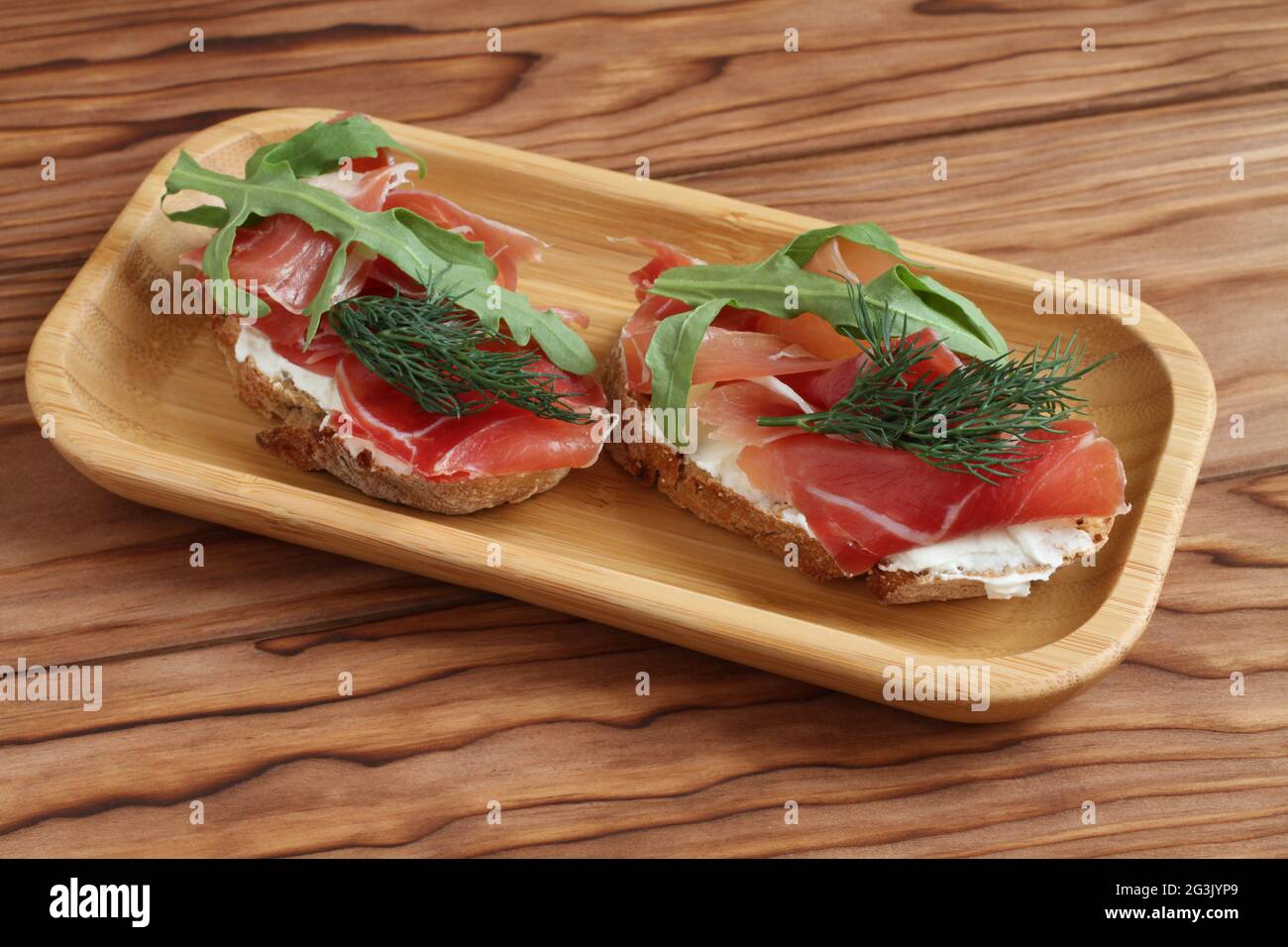 Buckwheat toasts with goat cheese, ham and arugula lying on a bamboo plate on a wooden table. Closeup Stock Photo