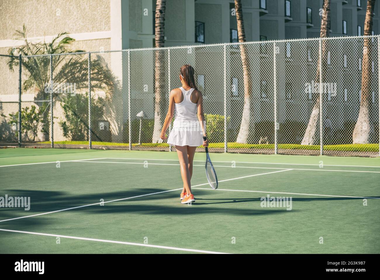 Tennis player woman playing on green hard court at resort hotel outside wearing white tennis dress outfit from behind. Stock Photo