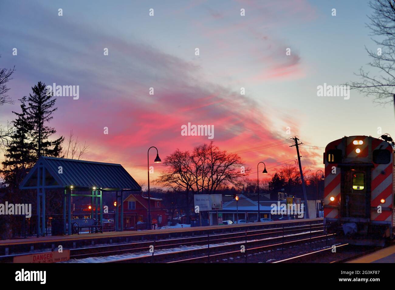 Winfield, Illinois, USA. The setting sun fills high clouds with color as a Metra communter train departs the railroad station on its continuing journe Stock Photo
