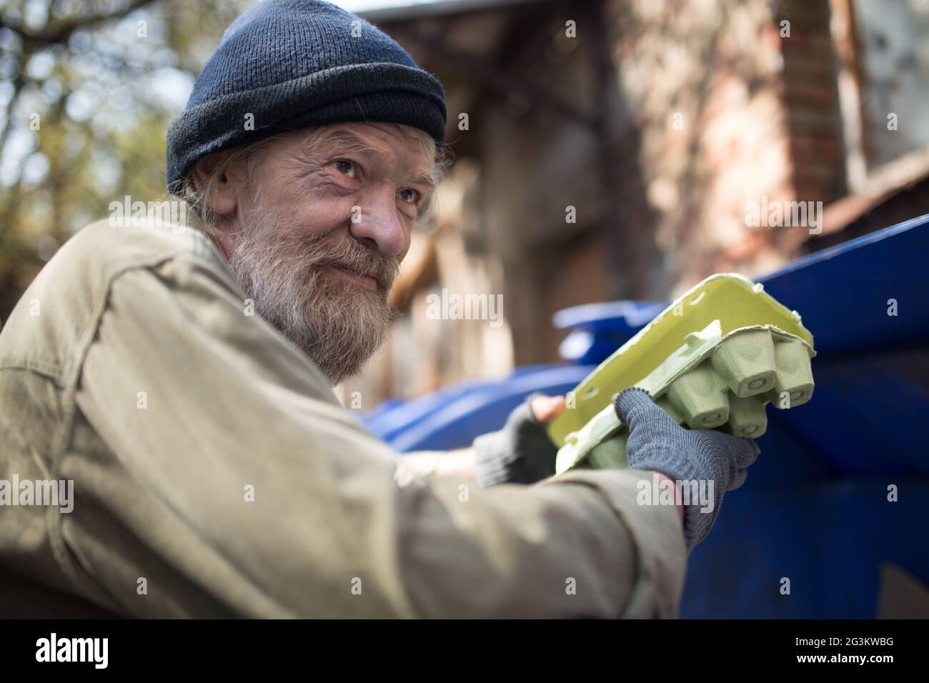 Dirty homeless man holding packing for eggs, standing by the trash can. Stock Photo