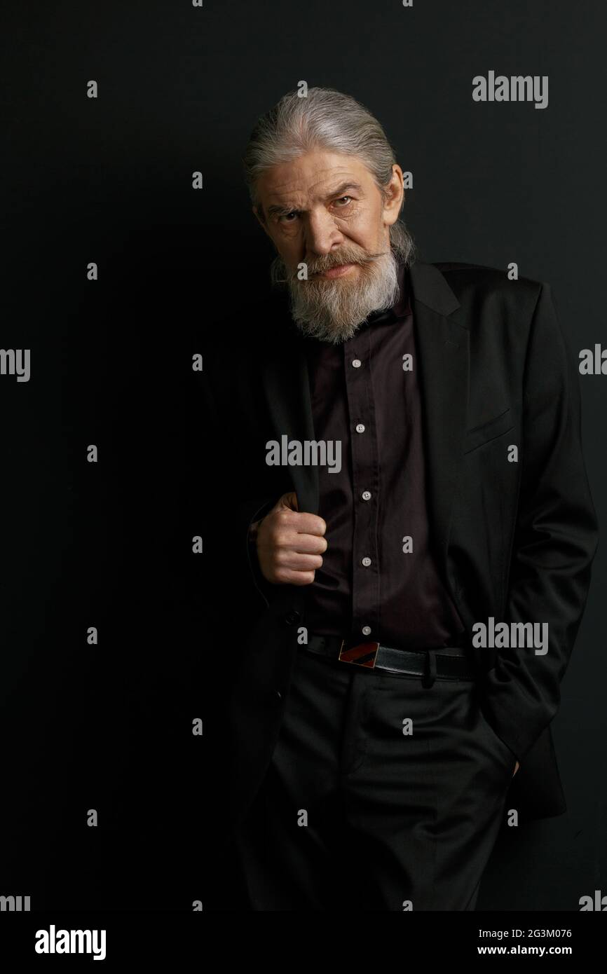 Stylish aged man with lonf hair and beard standing in studio against black wall. Stock Photo