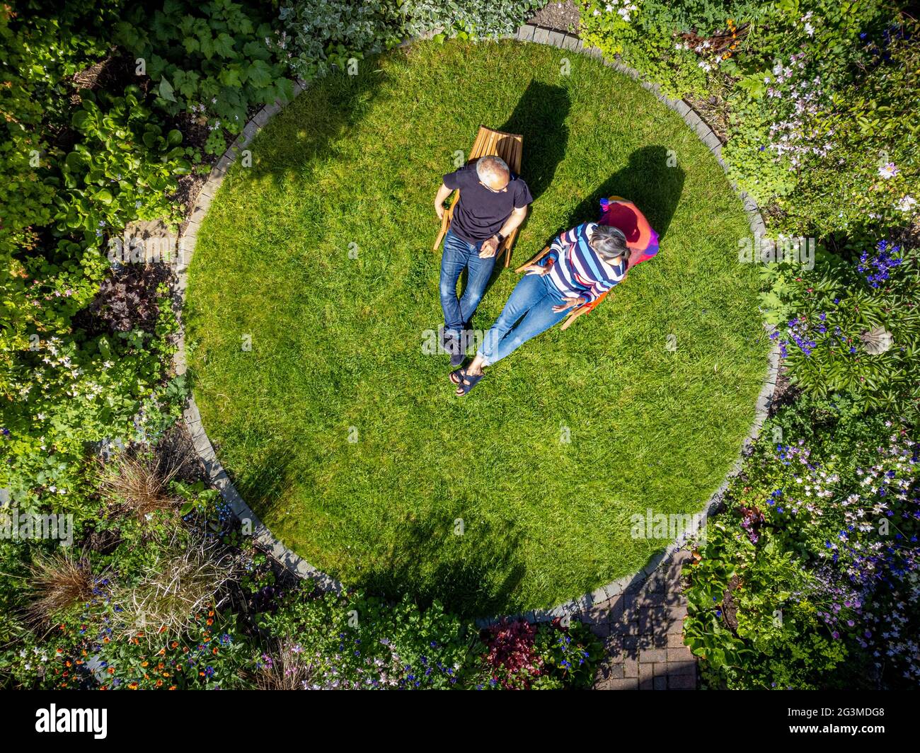 Aerial view of domestic garden with circular lawn and mature, casually dressed caucasian male and female sat on wooden deckchairs. Stock Photo