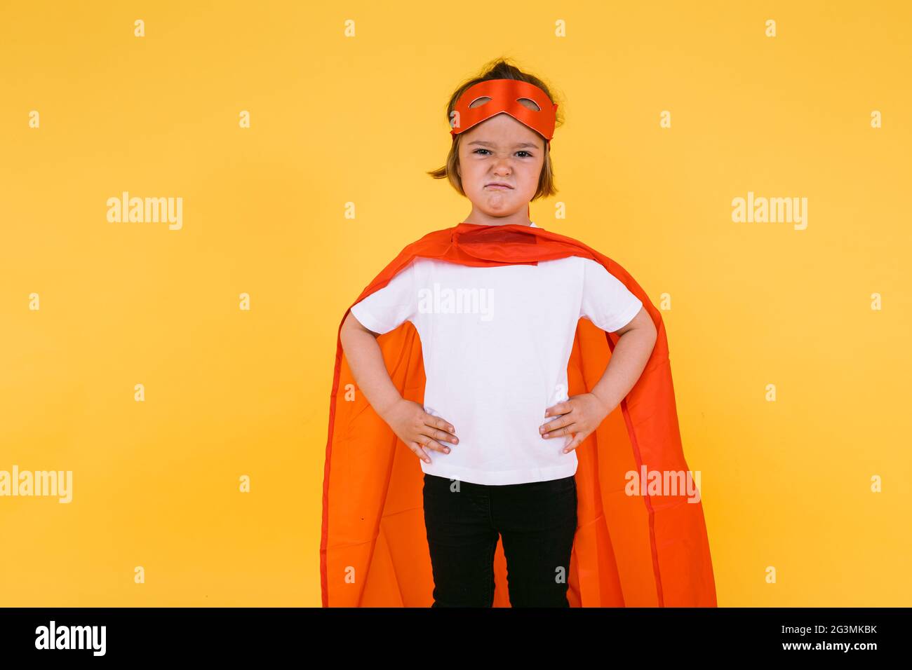 Little blonde girl dressed as a superheroine superhero with a cape and red mask, angry, with her arms akimbo serious, on yellow background Stock Photo