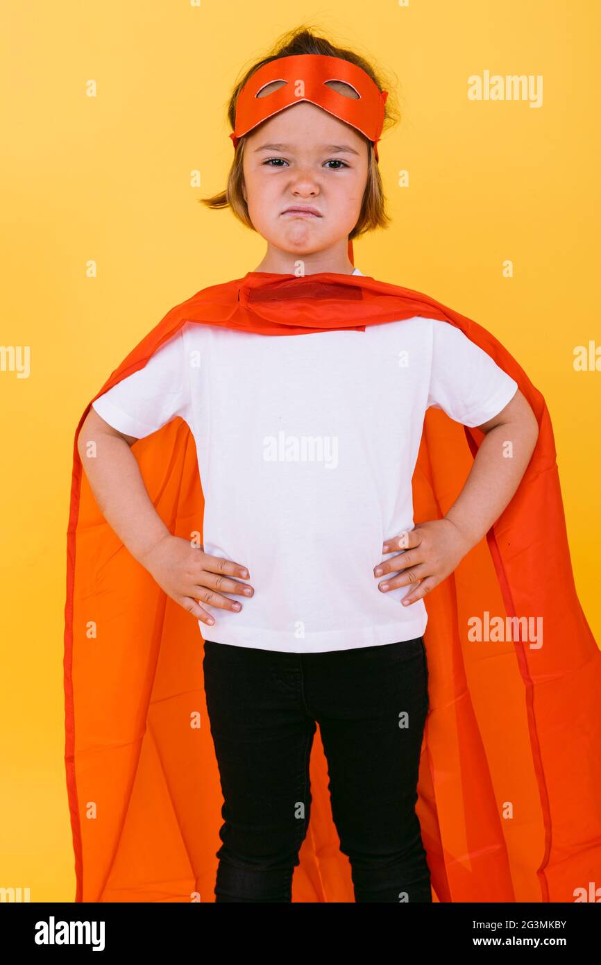 Little blonde girl dressed as a superheroine superhero with a cape and red mask, angry, with her arms akimbo serious, on yellow background Stock Photo