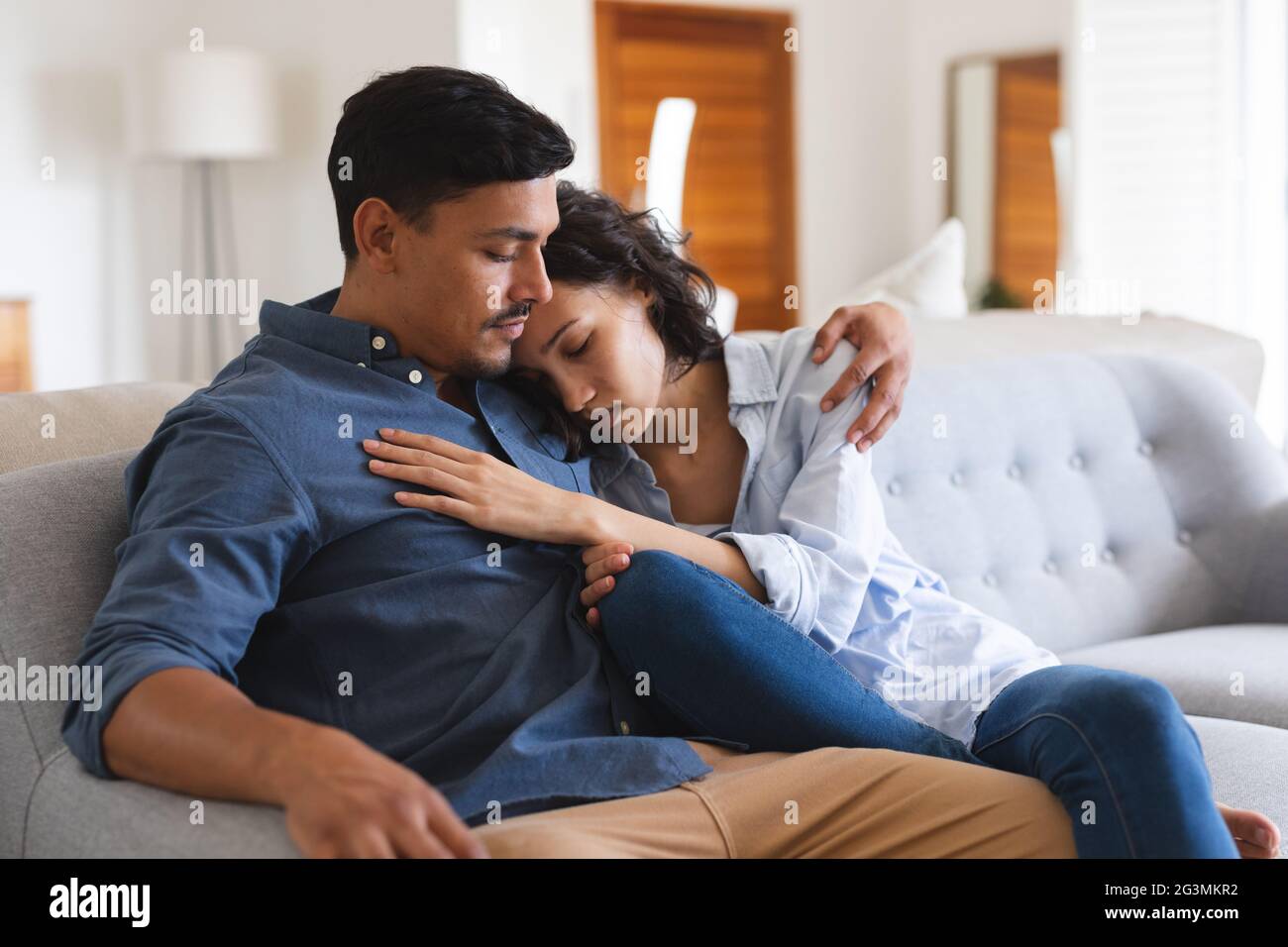 Sad hispanic couple sitting on couch in living room embracing Stock Photo