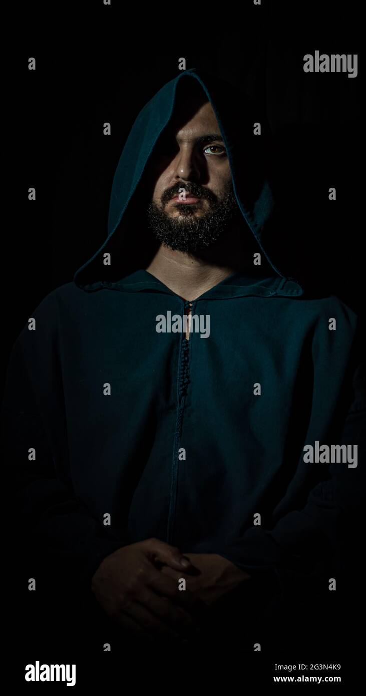 A african muslim man with a beard sits in a dark place looking at the camera and wears a blue traditional Moroccan djellaba. Stock Photo