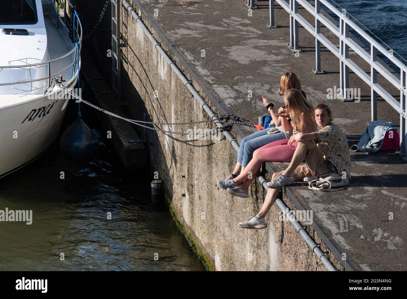 Two girls and a boy are sitting on the edge of a quay wall along the river Maas in Maastricht on a warm summer day and eating an ice cream Stock Photo