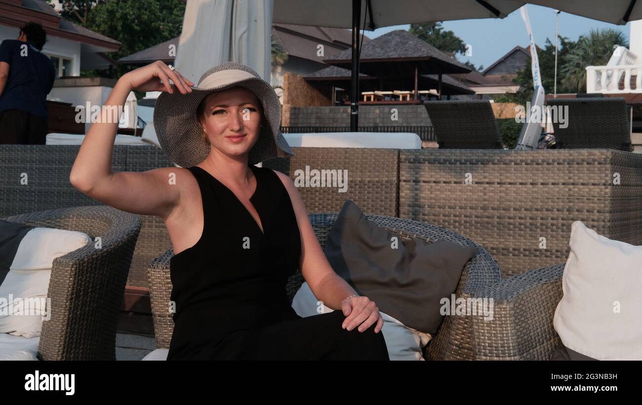 Eastern European Caucasian woman middle aged blonde hair light skin pale complexion in black dress gray straw hat outside summer time sitting Stock Photo