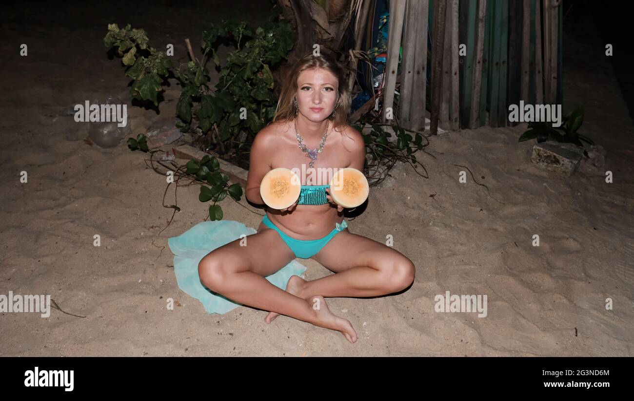 Caucasian woman middle aged blonde hair light skin pale complexion in Blue Bikini at night sitting on the sand wearing a neckless holding cantaloupe Stock Photo