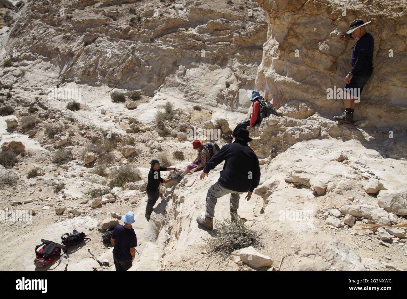 Hikers, senior adults in their 50s, 60s and 70s walk & slide down a high steep and dangerous rock to a ravine, one man helps the others, Negev Desert. Stock Photo