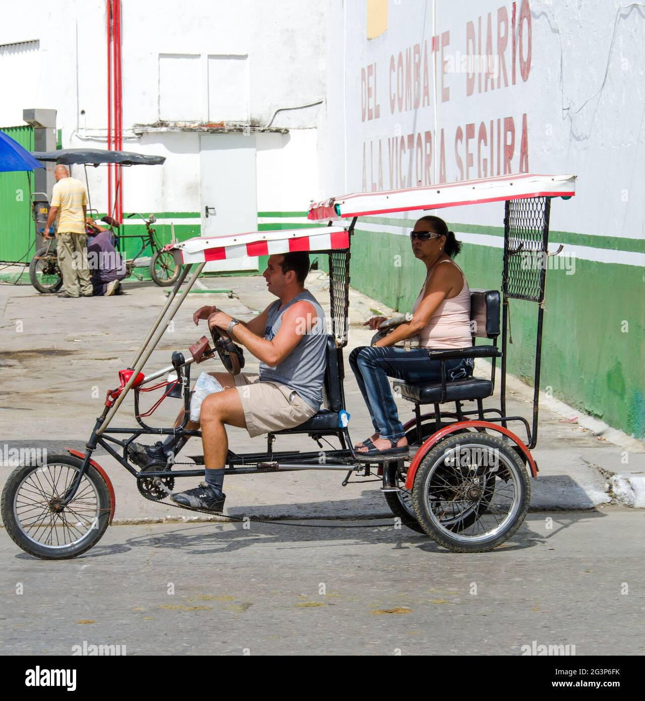 Two people ride a  bici taxi or bicitaxi in a Cuban street.  The man is pedaling the cart underits red striped awning, wearing a gray tank top, khaki Stock Photo