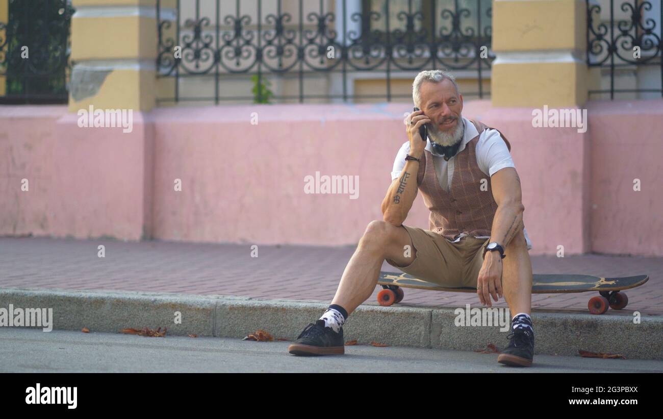 Middle-aged skater sits on longboard and speaks on mobile Stock Photo