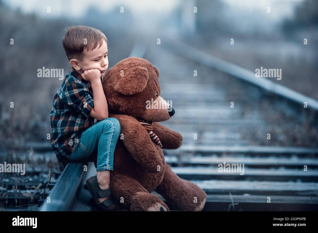 A little boy with a big teddy bear sits on the rails of the railway ...