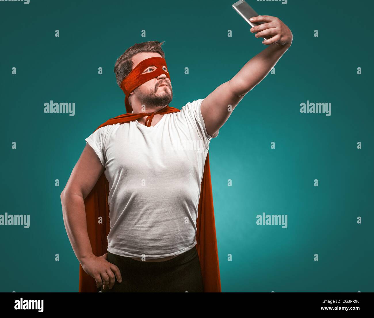 Super Hero man Takes Selfie By Mobile Phones. Man In A Super Hero Costume In A Red Mask And Cloak Isolated On Biscay Green Backg Stock Photo