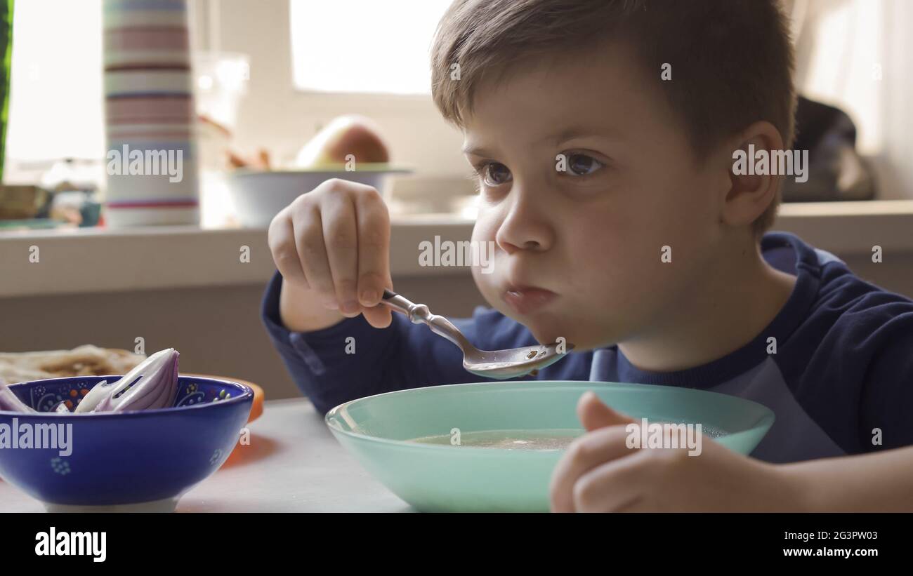 Hungry preschool child eating soup from a bowl. A serious boy scored a mouthful of soup sitting with big cheeks. Good appetite c Stock Photo
