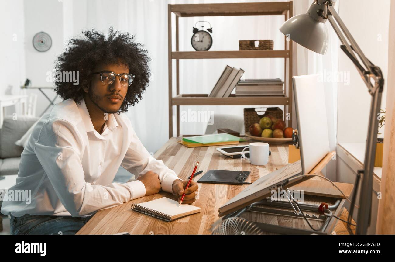 Creative man writes in Note Pad working laptop at Home work place. Arabic business or student in eyeglasses looking at camera. F Stock Photo