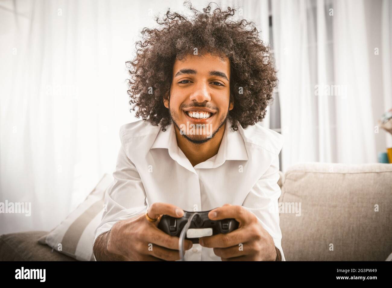 Arabic gay playing computer game. Young man holds Play Station joystick sitting at sofa in backlit. Smiling handsome looking at Stock Photo