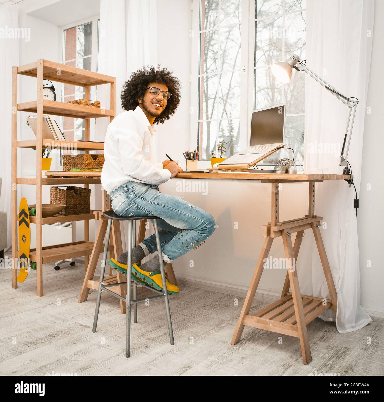 Smart millennial working laptop at home sitting near window. Arabic freelancer in white shirt and jeans smiles looking at camera Stock Photo