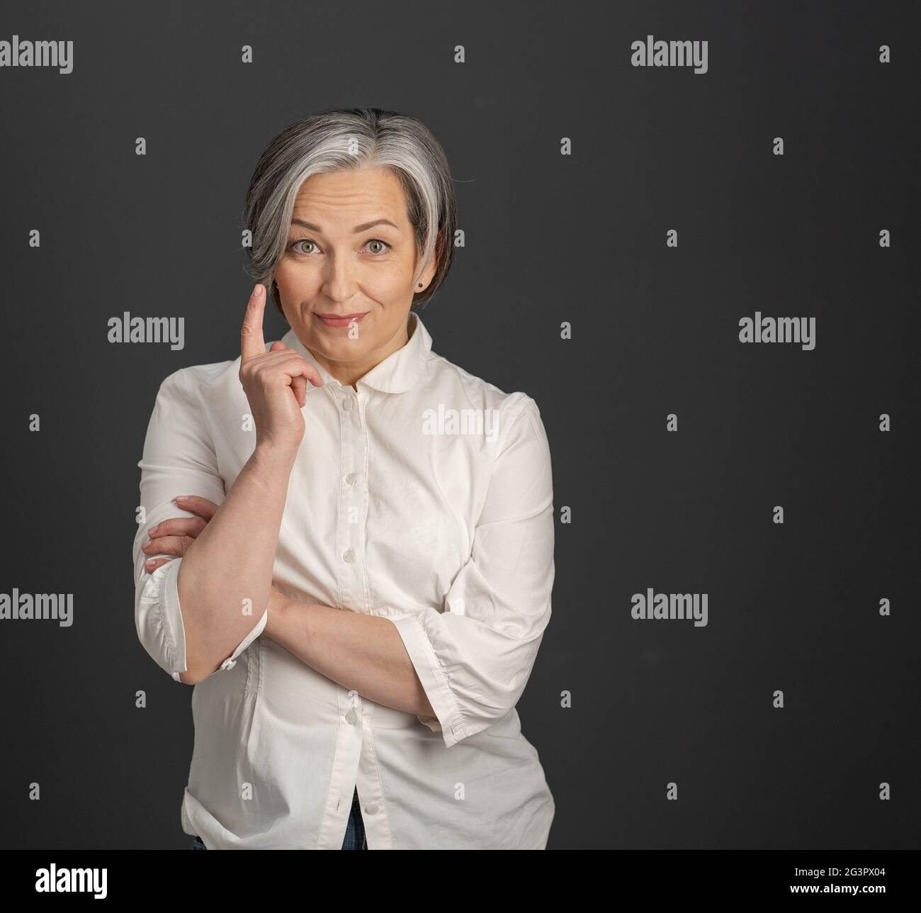 Have idea gesture from mature woman. Gray-haired businesswoman raised her index finger up while looking at camera posing on grey Stock Photo