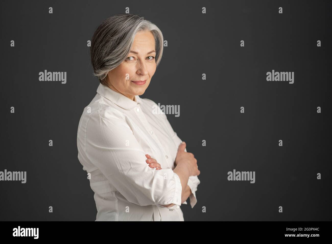 Pretty white-haired woman smiles looking at camera with arms crossed. Stylish caucasian businesswoman isolated on dark gray back Stock Photo