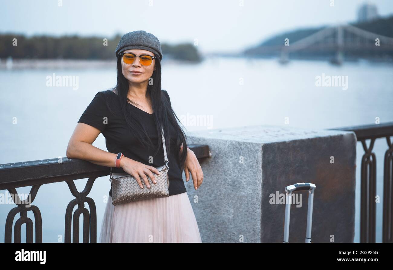 Female traveler in sunglasses resting on embankment of the river. Beautiful asian woman with suitcase travels in european city. Stock Photo