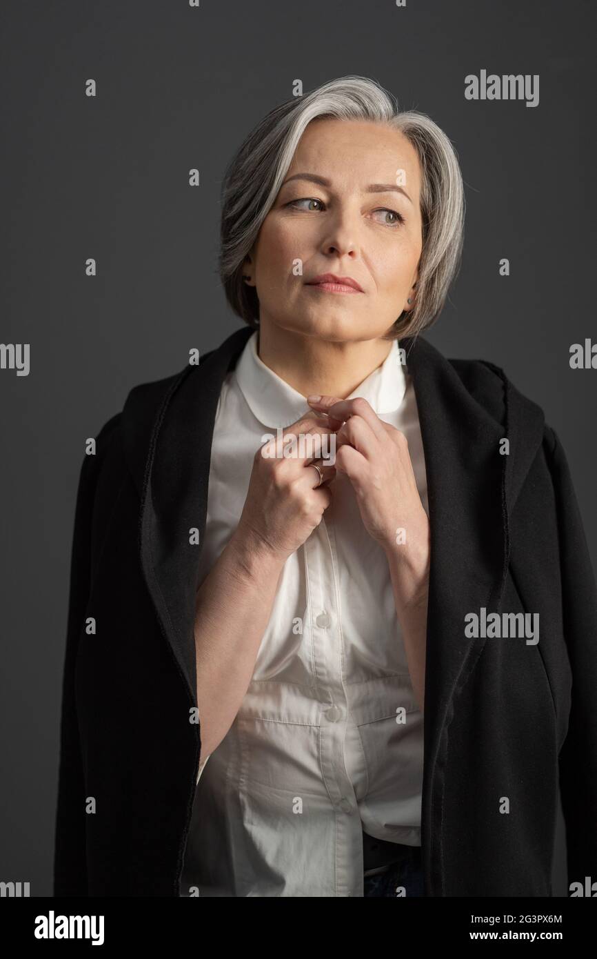 Charming gray-haired woman thoughtfully looks away adjusting collar of white shirt, throwing black jacket over her shoulders. Is Stock Photo