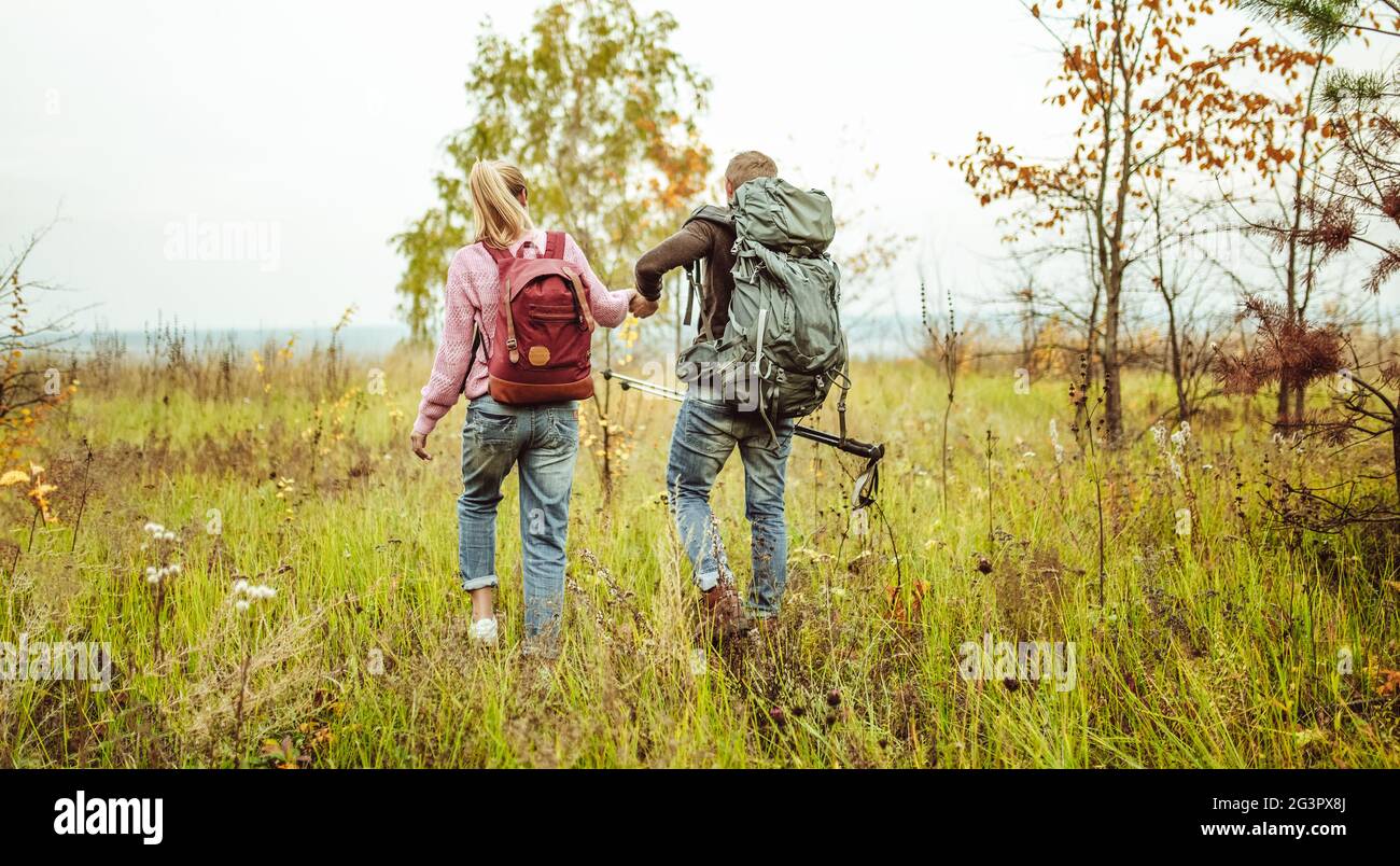 Rear view of a traveling couple of backpacker wading outdoors through an autumn field holding hands with hiking poles. Hiking co Stock Photo