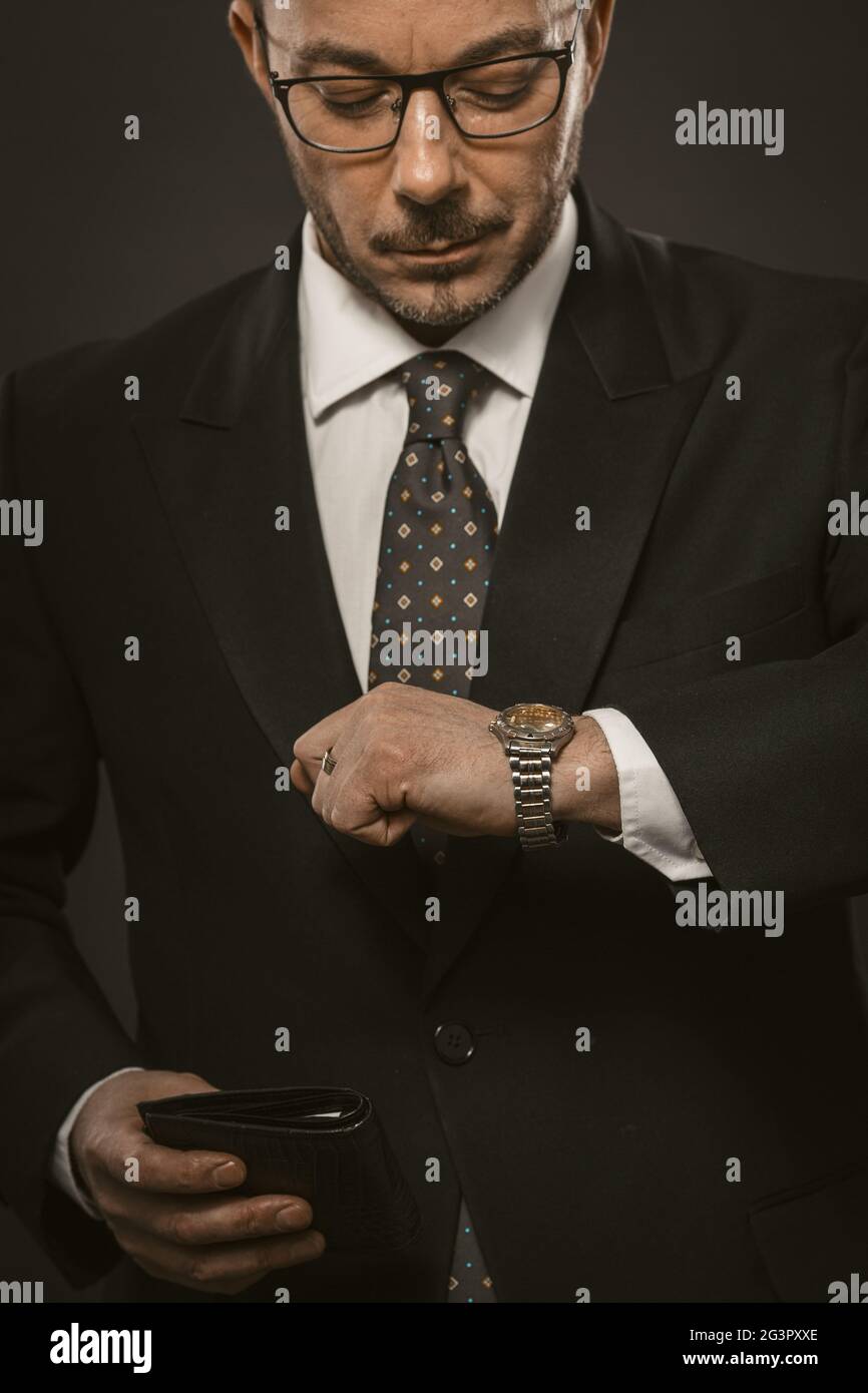 Punctual businessman checks time looking on his wristwatch while holding wallet in other hand. Well dressed bearded man in eyegl Stock Photo