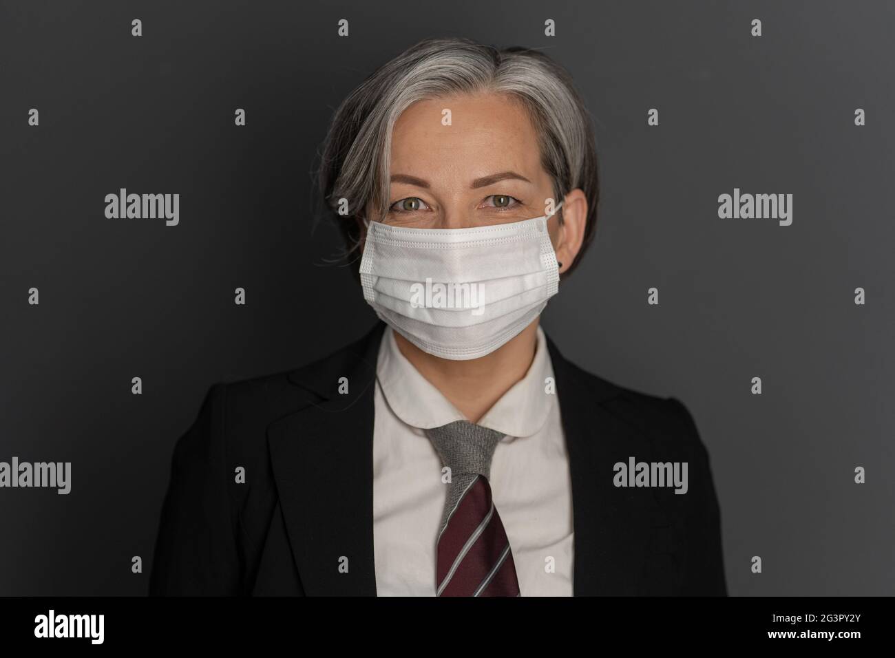Pretty Caucasian businesswoman wearing protective mask and formalwear smiles on gray background. Copy space on both sides. Close Stock Photo