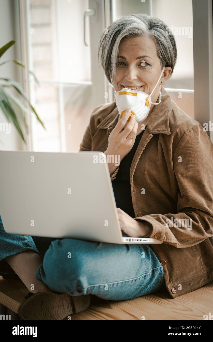 Grey haired woman removes mask affably looking at camera. Caucasian lady uses computer sitting