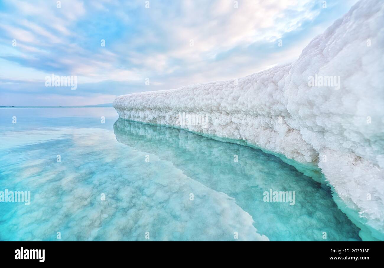 Sand completely covered with crystalline salt looks like ice or snow on shore of Dead Sea, turquoise blue water near, sky colored with morning sun dis Stock Photo