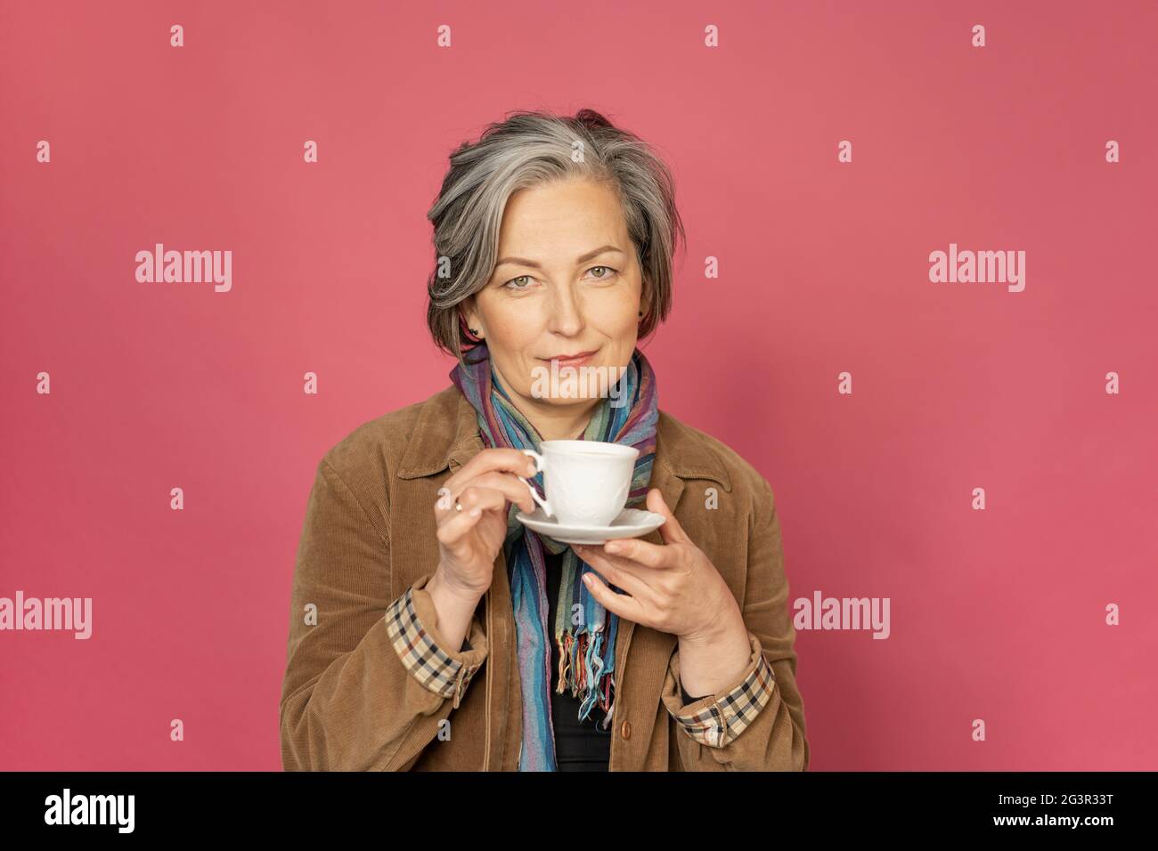 Charming Caucasian woman drinks coffee holding white cup. Pretty mature lady smiles slightly isolated on pink background. Beauty Stock Photo