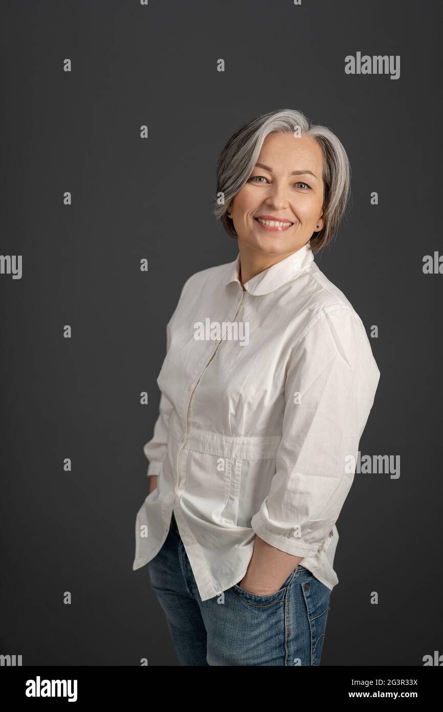 Toothy smiling mature woman stands hands in pockets looking at camera. Gray-haired woman in jeans and white blouse posing on gra Stock Photo