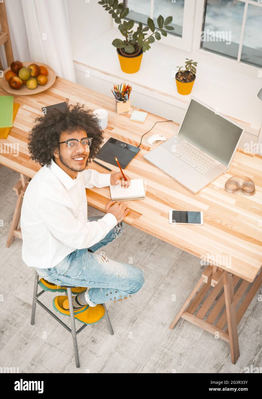 Creative millennial man working at home workplace. Young Arab businessman looks at camera sitting at wooden desk with computer, Stock Photo