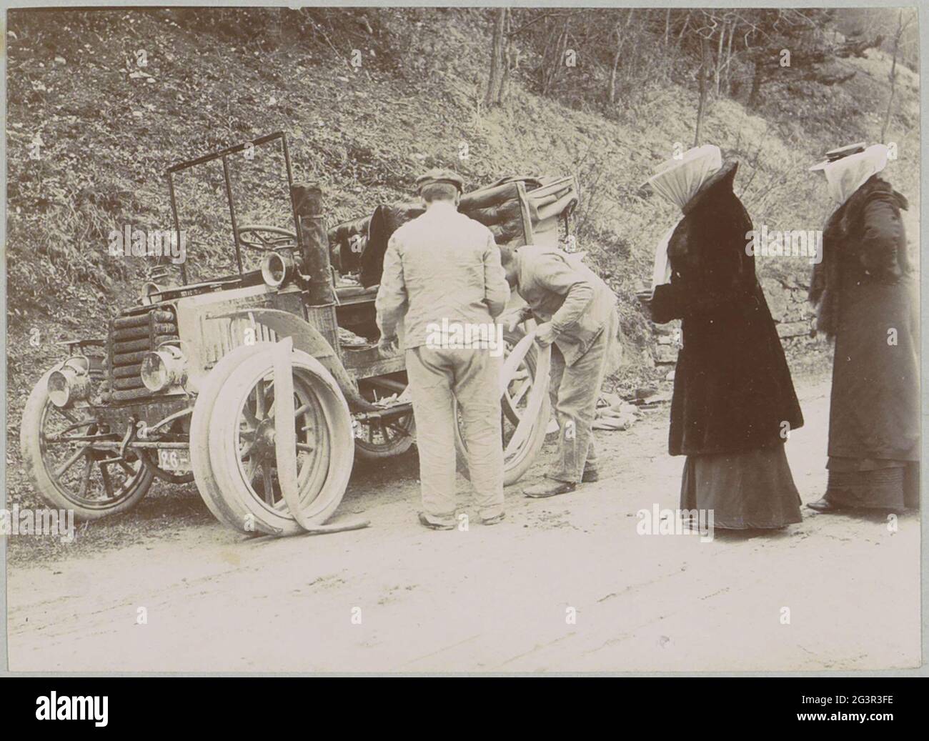 Companation looks at automobile tire change, presumably in the French Alps. Part of photo album from a French amateur photographer with recordings of trips in France, Spain, Belgium, Luxembourg and the Netherlands, the first automobiles and autoraces. Stock Photo