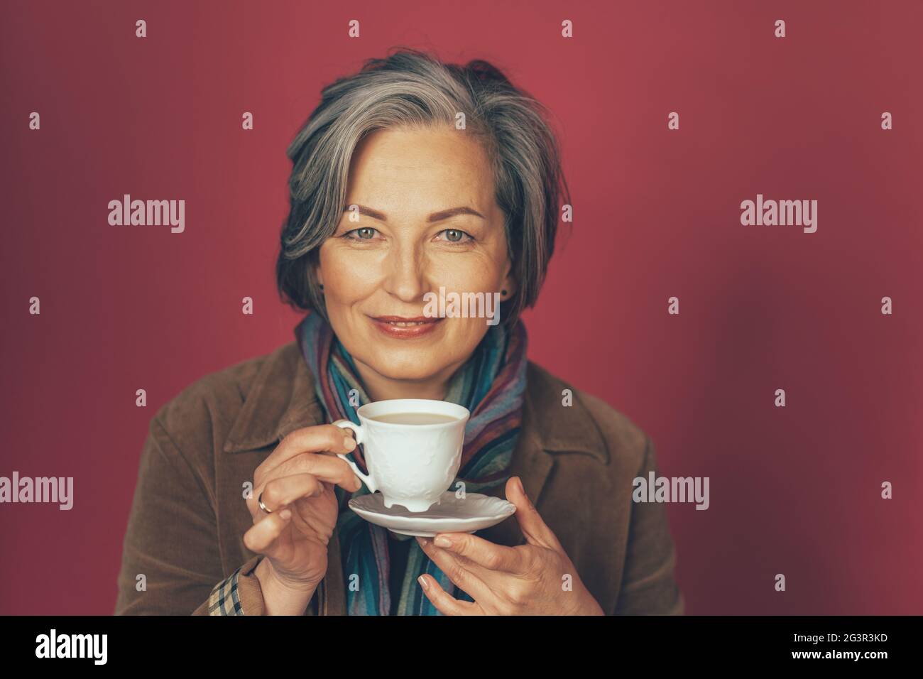Beautiful caucasian woman holds white cup drinking coffee in studio on pink background. Happy gray-haired female looking at came Stock Photo