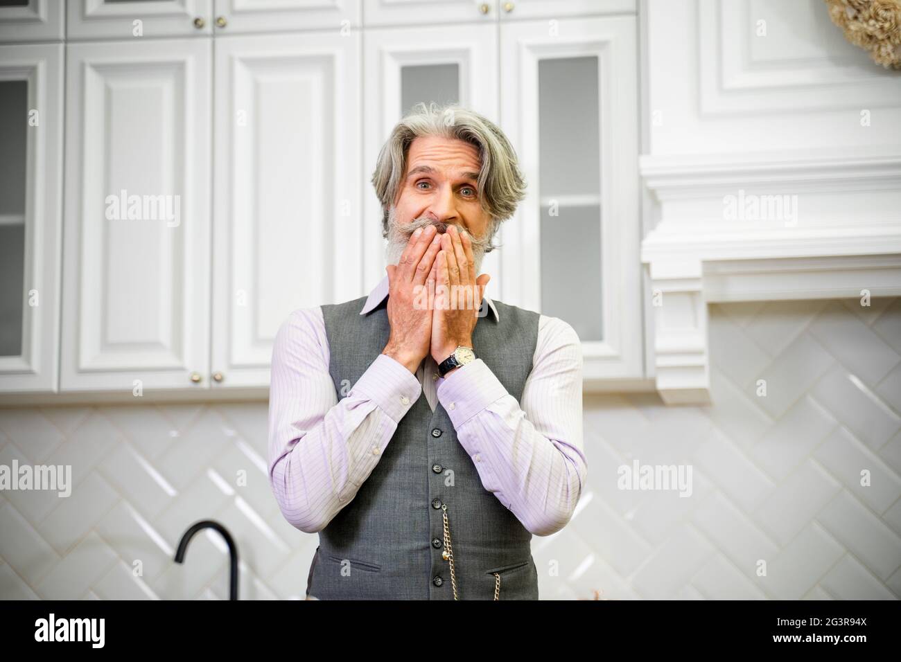 Elegant senior grey haired grandfather in formal wear being surprised when receiving present for his birthday, standing in stylish kitchen at home holding hands over mouth with excited expression Stock Photo