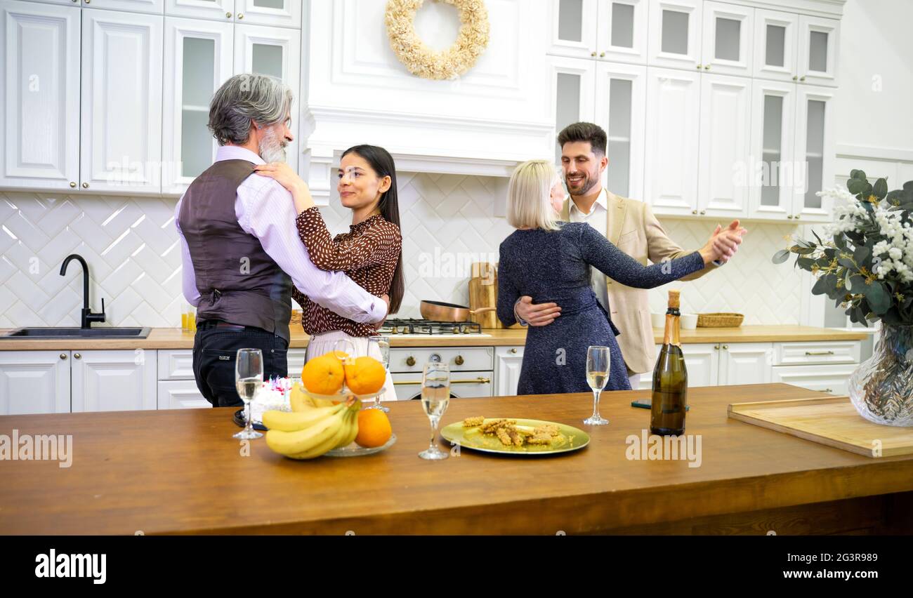 Happy family members dancing during birthday celebration in kitchen near table with cake and champagne at home, grandparents and young married couple having fun and enjoying home party Stock Photo