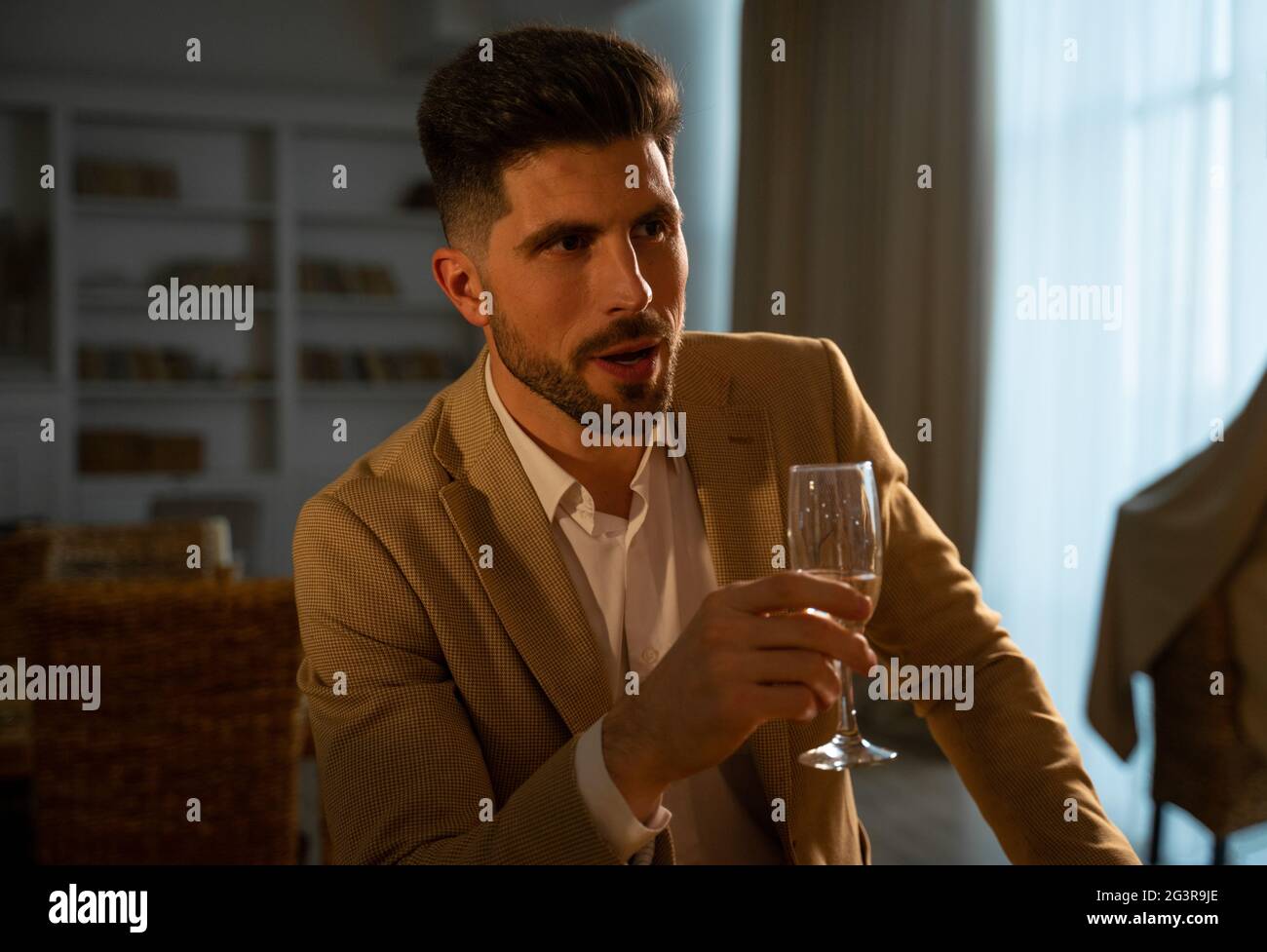 Handsome young man in formal festive suit raising his glass of champagne while making a toast in stylish living room at holiday dinner in evening, wishing happy birthday. Celebration concept Stock Photo