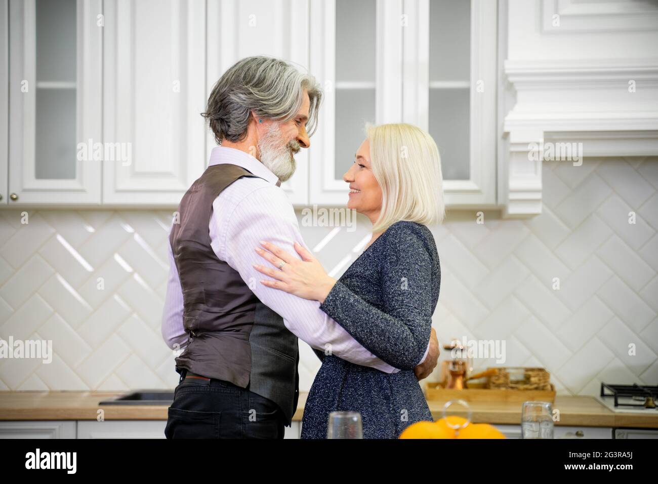 Senior married couple in festive wear dancing in stylish kitchen near table with champagne at home during birthday celebration. Smiling husband looking at wife and expressing his love. Family concept Stock Photo