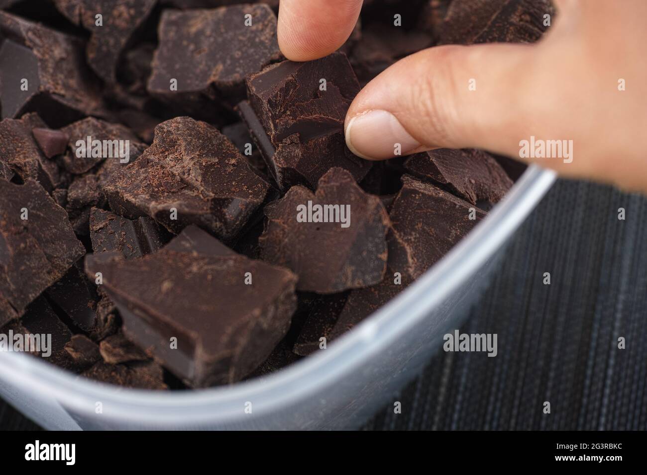 A woman taking a piece of broken organic homemade dark chocolate out of a plastic container. Close up. Stock Photo