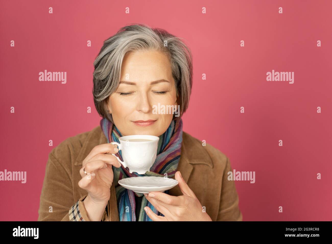 Charming adult woman drinks coffee holding white cup and closed eyes. Isolated on pink background. Studio shot. rest concept Stock Photo