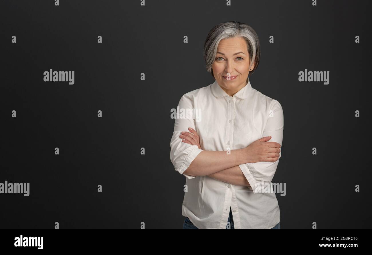 Graying pretty woman in white shirt stands arms-crossed isolated on dark gray background. High quality photo Stock Photo