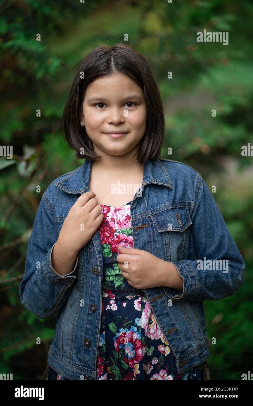 Cute little girl wearing plaid dress and denim jacket posing against backdrop of green nature Stock Photo