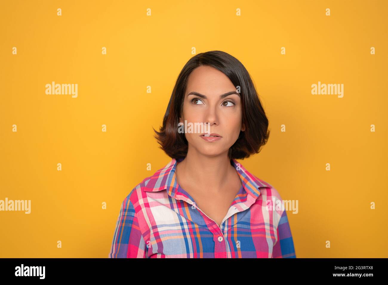 Thinking young woman makes serious decision. Pretty brunette looks up at copy space on left. Isolated on yellow background Stock Photo