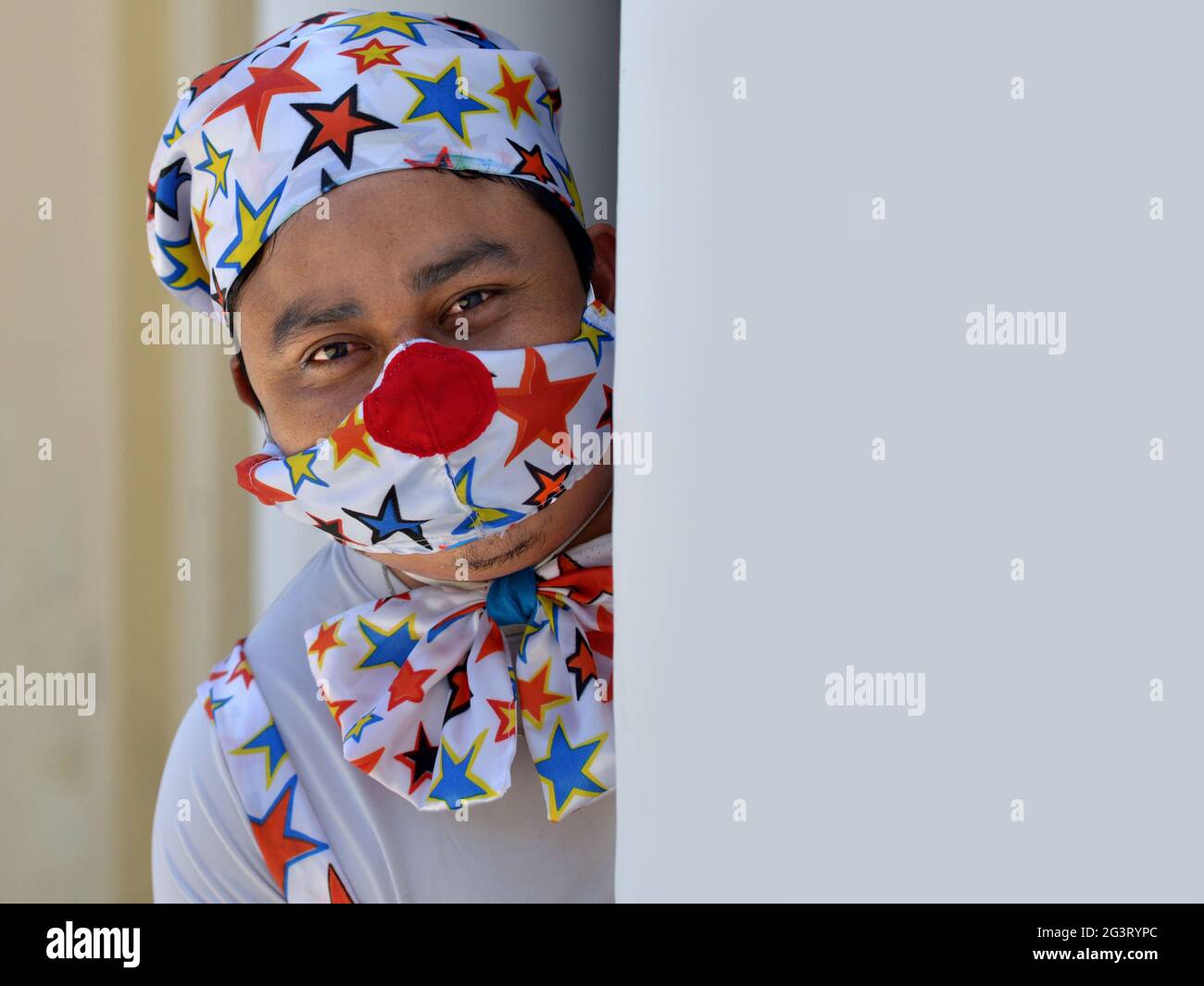 Funny male clown wears star pattern clown outfit and star pattern fabric face mask and peeks around a corner during the global coronavirus pandemic. Stock Photo
